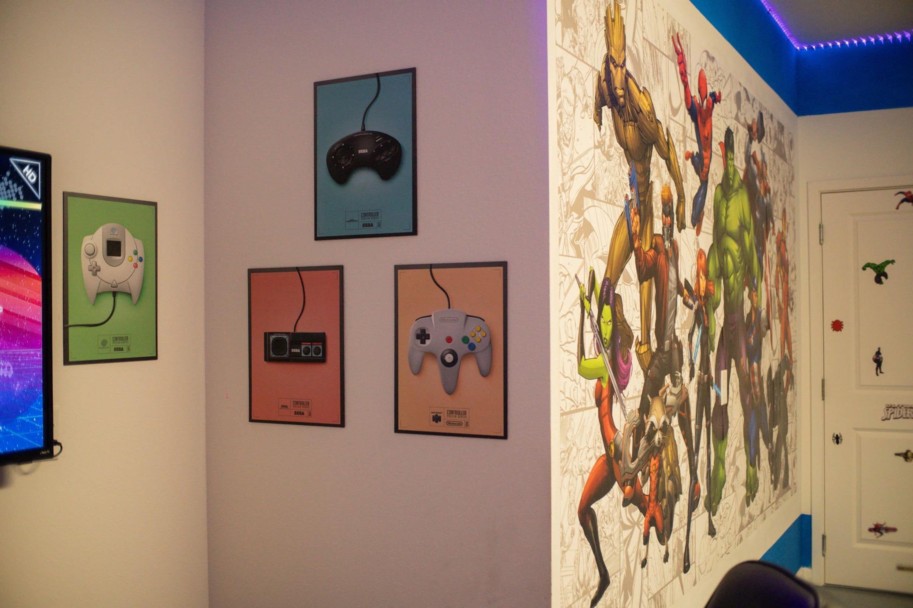Una pared decorada con controles de videojuegos enmarcados y un gran mural de superhéroes.