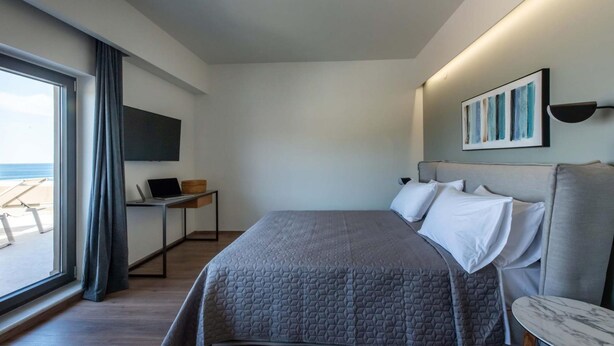 A modern hotel room with a large bed, a desk with a laptop, a TV mounted on the wall, and a view of the beach. - Lusol Seafront Villa Ii-free Heated Pool 100m Sea (Chania)