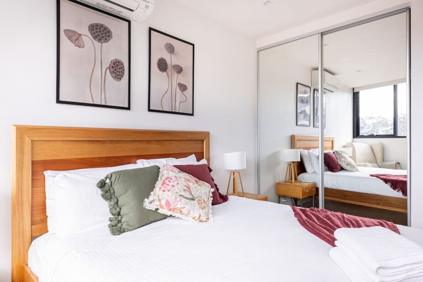 A hotel room with a wooden headboard, a large bed with white bedding, a nightstand with a lamp, and a large mirror reflecting the room.
