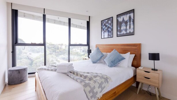 A modern bedroom with a large bed, two bedside tables, and a view of a bridge through large windows. - Sanctuary-Effortless City Escape (Abbotsford)