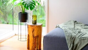 A room with a large window, a wooden side table with a plant, a blue sofa, and a green throw blanket. - Retreat, short drive to Beach (Rye)