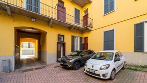 Ein Innenhof mit zwei parkenden Autos, einem gelben Gebäude mit Balkonen und einer Tür, die zu einem Innenraum führt.