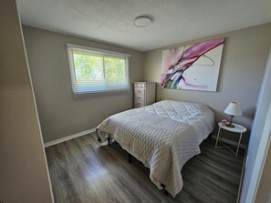 Quarto com cama, penteadeira, janela com cortinas, quadro na parede e mesa de cabeceira com abajur.