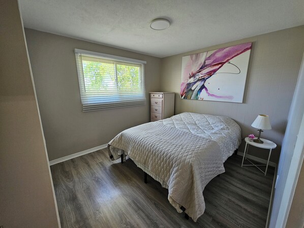 A bedroom with a bed, a dresser, a window with blinds, a painting on the wall, and a nightstand with a lamp.