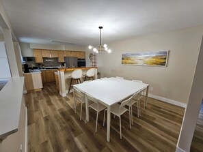 Uma cozinha moderna com armários de madeira, uma área de jantar com mesa e cadeiras brancas, e um quadro na parede.