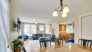 Uma sala de estar espaçosa com uma grande mesa de jantar de madeira, sofás cinzas e uma cozinha ao fundo.