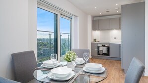 Un espace repas moderne avec une table en verre, des chaises recouvertes d’un tissu gris et une vue sur la ville grâce à de grandes fenêtres.