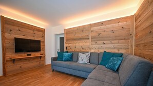 Ein modernes Wohnzimmer mit einem grauen Ecksofa, Holzwandverkleidung und einem an einer Holzablage montierten Flachbildfernseher.