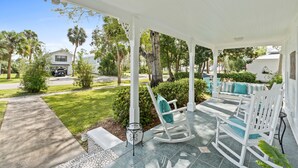 A covered porch with white rocking chairs and a small table, overlooking a well-maintained lawn and neighboring houses. - Bay view Historic home walking to St. Joseph Bay and Downtown.  (Port St. Joe)