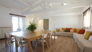 Un vaste espace de vie comprenant une table à manger en bois, des chaises blanches, un canapé agrémenté de coussins colorés et une plante en pot.