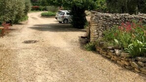 Une allée de gravier menant à une voiture garée, entourée de verdure et d’un mur de pierres.