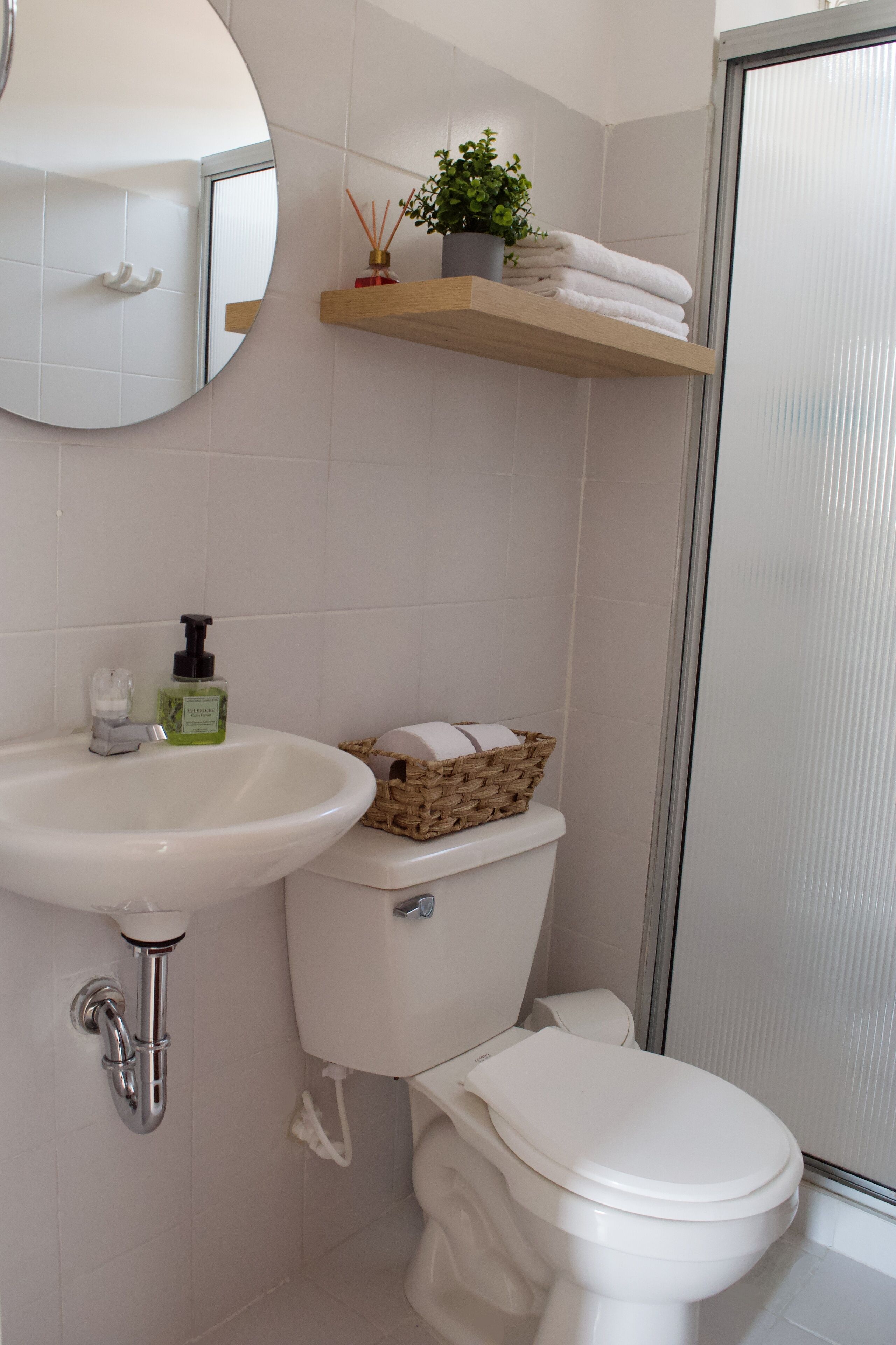 Ein Badezimmer mit einem weißen Waschbecken, einer Toilette und einem Spiegel. Ein Regal zeigt eine Pflanze und Handtücher.