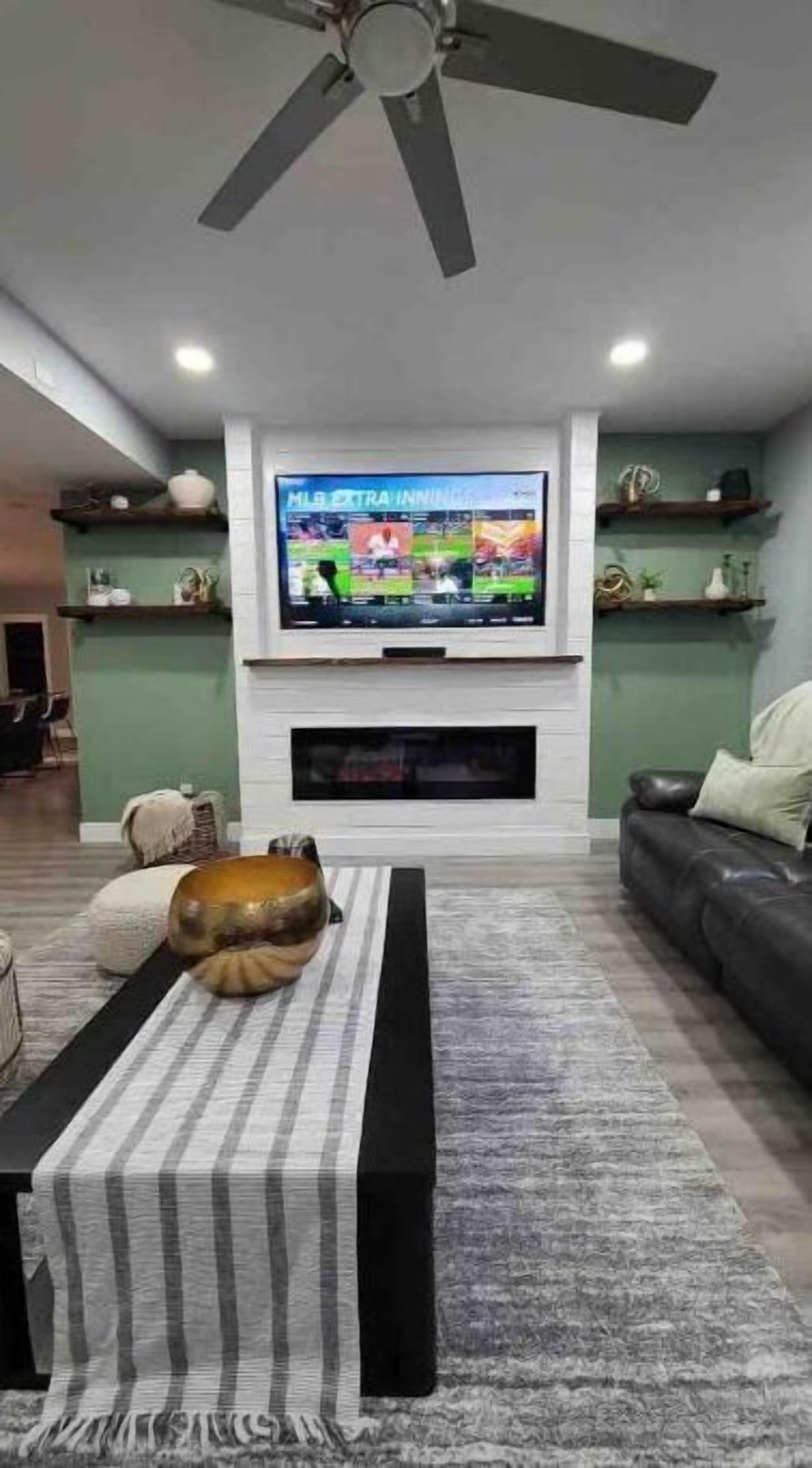 A living room with a flat-screen TV mounted above a fireplace, a ceiling fan, and a sofa.