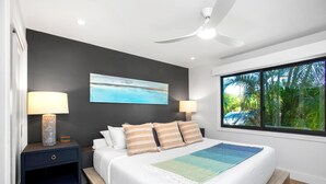 Un dormitorio con cama, dos mesitas de noche, ventilador de techo, un cuadro y ventana con vista a palmeras.