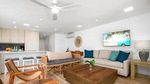 Una sala de estar moderna con sofá, mesa de centro y sillas, una zona de cocina con barra y un ventilador de techo.