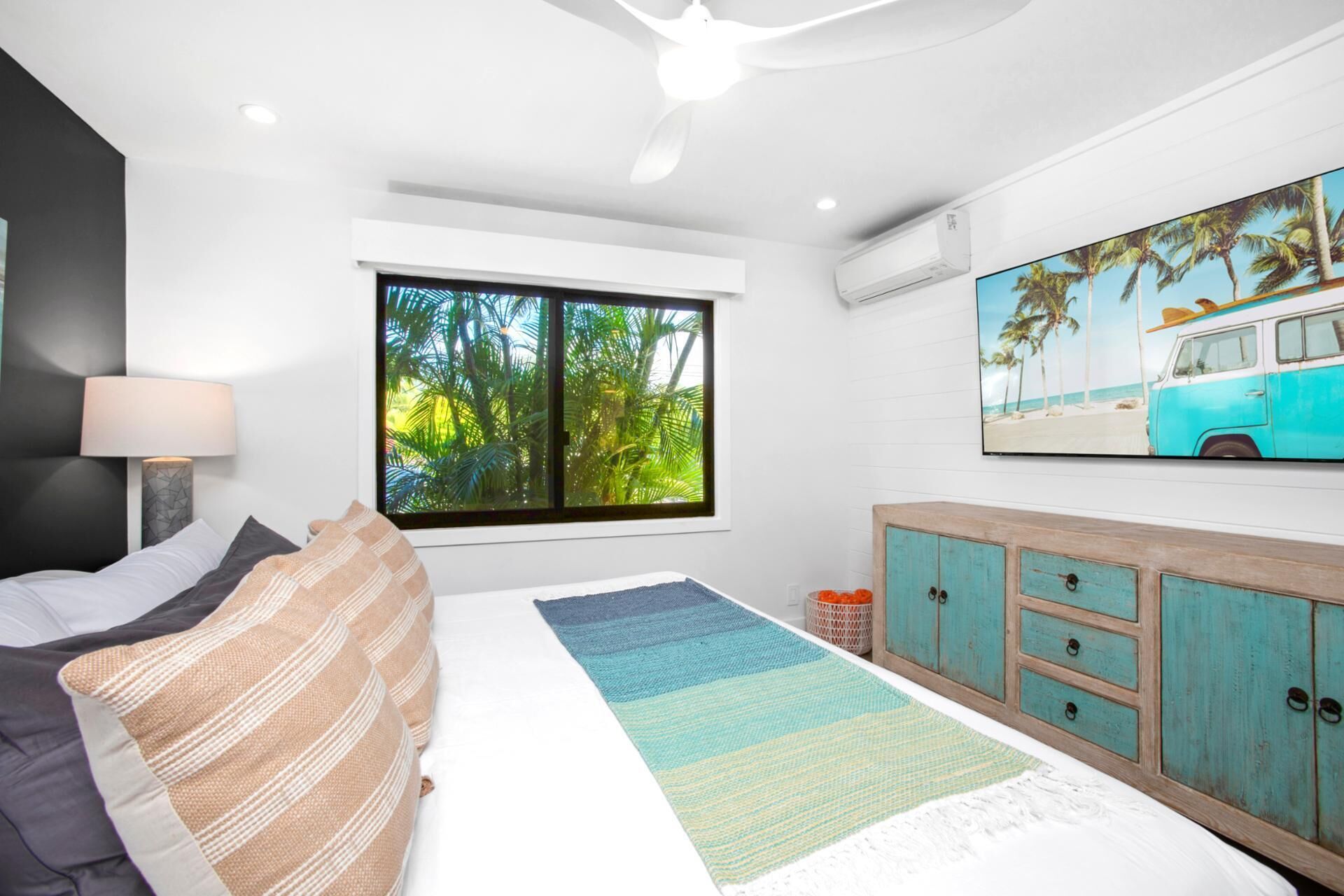 A bedroom with a bed, a window with a view of palm trees, a ceiling fan, and a wall-mounted TV.