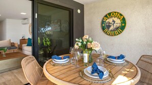 Un comedor con una mesa redonda de madera dispuesta para cuatro personas, un letrero en la pared que dice "Aloha Hawaii" y una puerta de vidrio que da a un espacio de estar.