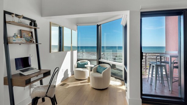 A modern living room with a view of the beach, a balcony with outdoor furniture, and a desk with a laptop.