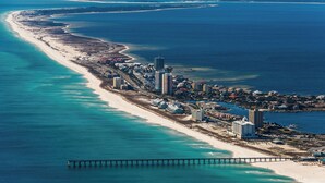 An aerial view of a coastal resort area with a long sandy beach, a pier extending into the ocean, and a cluster of buildings along the shore.