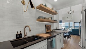 A modern kitchen with white subway tiles, a black countertop, and a stainless steel refrigerator. There are wooden shelves above the sink and a dining area with a view of the ocean.
