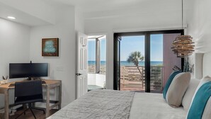 A bedroom with a bed, a desk with a computer, a TV, and a view of the beach.