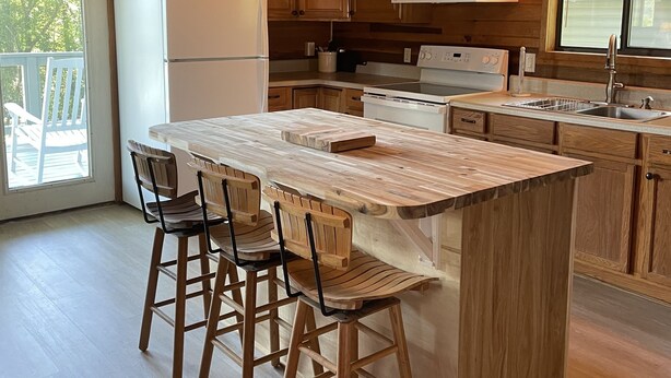 Cocina de cabaña de madera con una isla grande, taburetes de bar de madera, un refrigerador, una estufa y una ventana con vista a la vegetación.