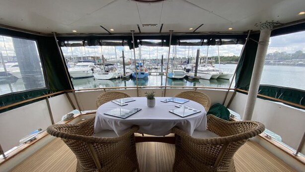 Ein Essbereich mit einem runden Tisch, Korbstühlen und Blick auf Boote in einer Marina.