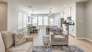 A spacious living room with a sofa, a coffee table, and a rug. There is a dining area with a table and chairs, and a kitchen with cabinets and appliances.