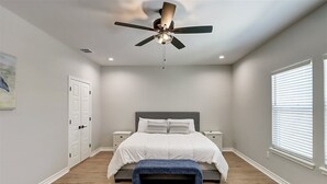 A bedroom with a bed, a ceiling fan, a bench, a nightstand, a window, and a door.