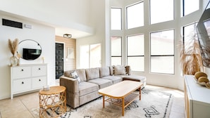 Uma sala de estar moderna com um sofá modular, uma mesa de centro e uma mesa lateral de vime redonda. Há um armário branco com gavetas e uma televisão de tela plana fixada na parede. O cômodo possui janelas amplas que permitem a entrada de luz natural e oferecem vista para o exterior.