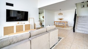 Uma sala de estar moderna com uma televisão de tela plana grande, um sofá seccional bege, uma área de jantar com mesa e cadeiras de madeira, e uma escadaria que leva a outro andar.