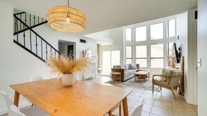 Uma sala ampla com uma mesa de jantar de madeira, cadeiras brancas, um sofá e uma poltrona. Há uma escada com guarda-corrimão preto e um candeeiro de vime suspenso.