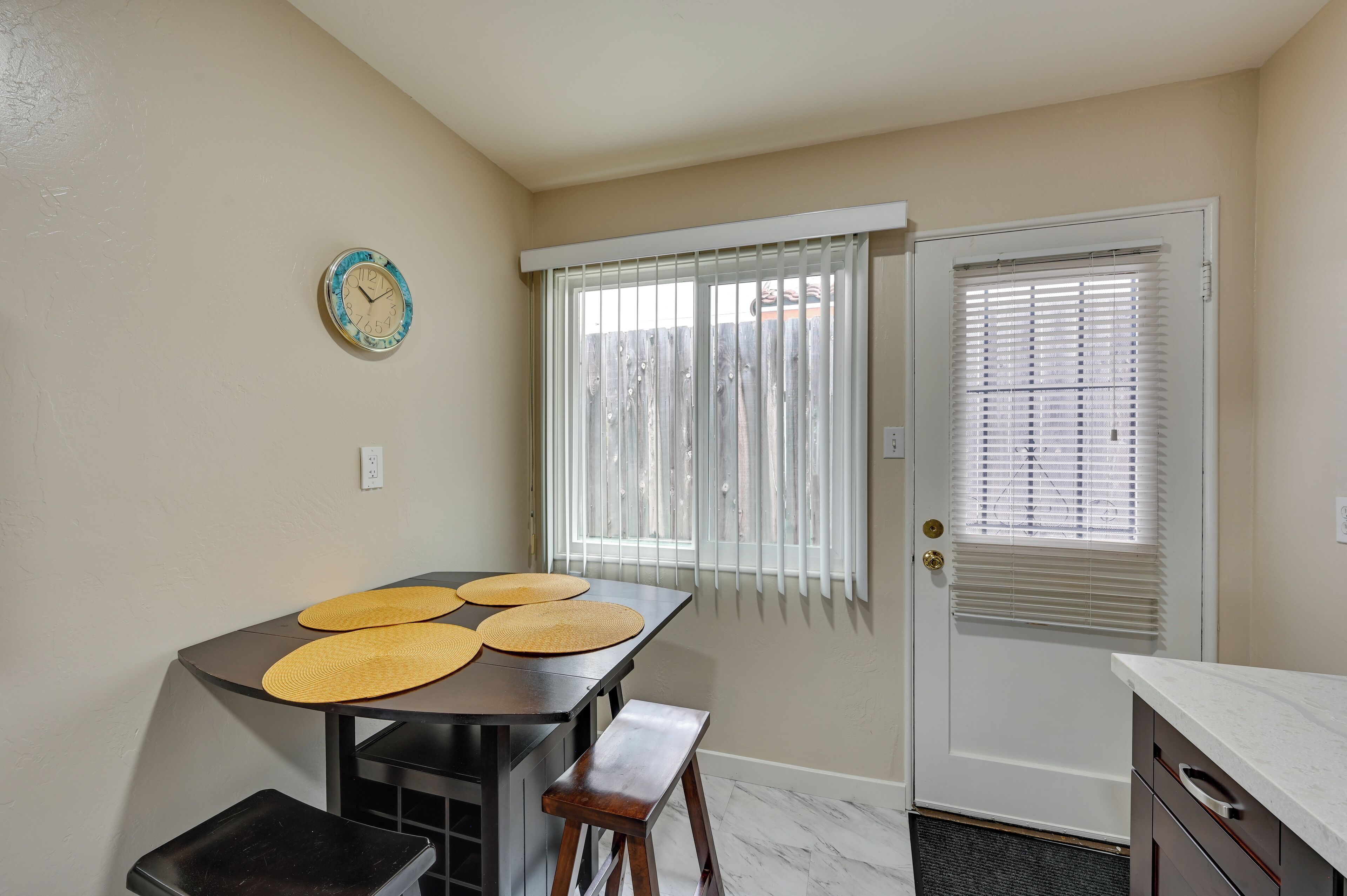 A compact kitchen with a dining table, bench seating, a clock on the wall, and a door with a window with blinds.