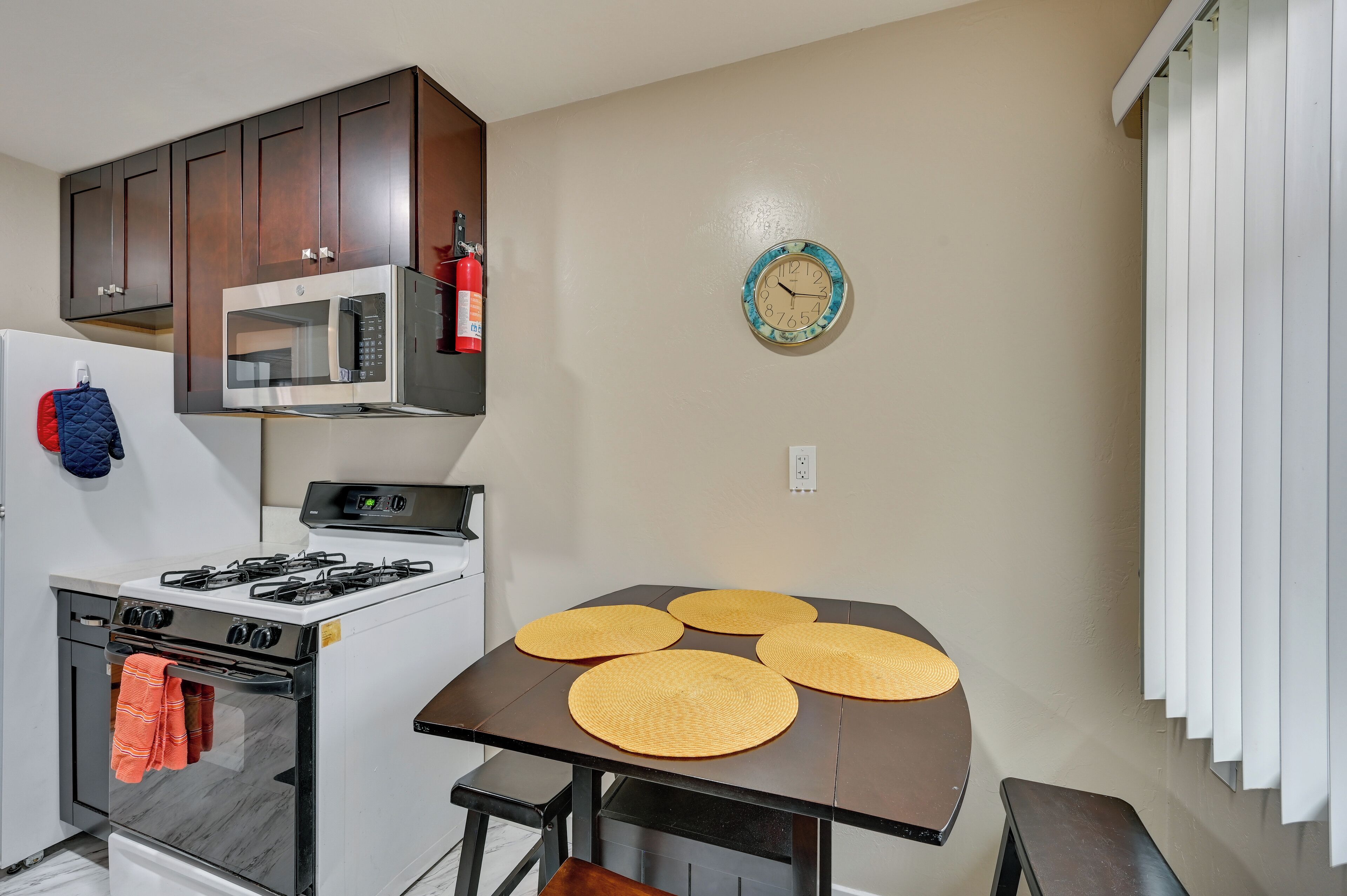 A kitchen with a stove, microwave, and cabinets. There is a clock on the wall and a table with placemats.