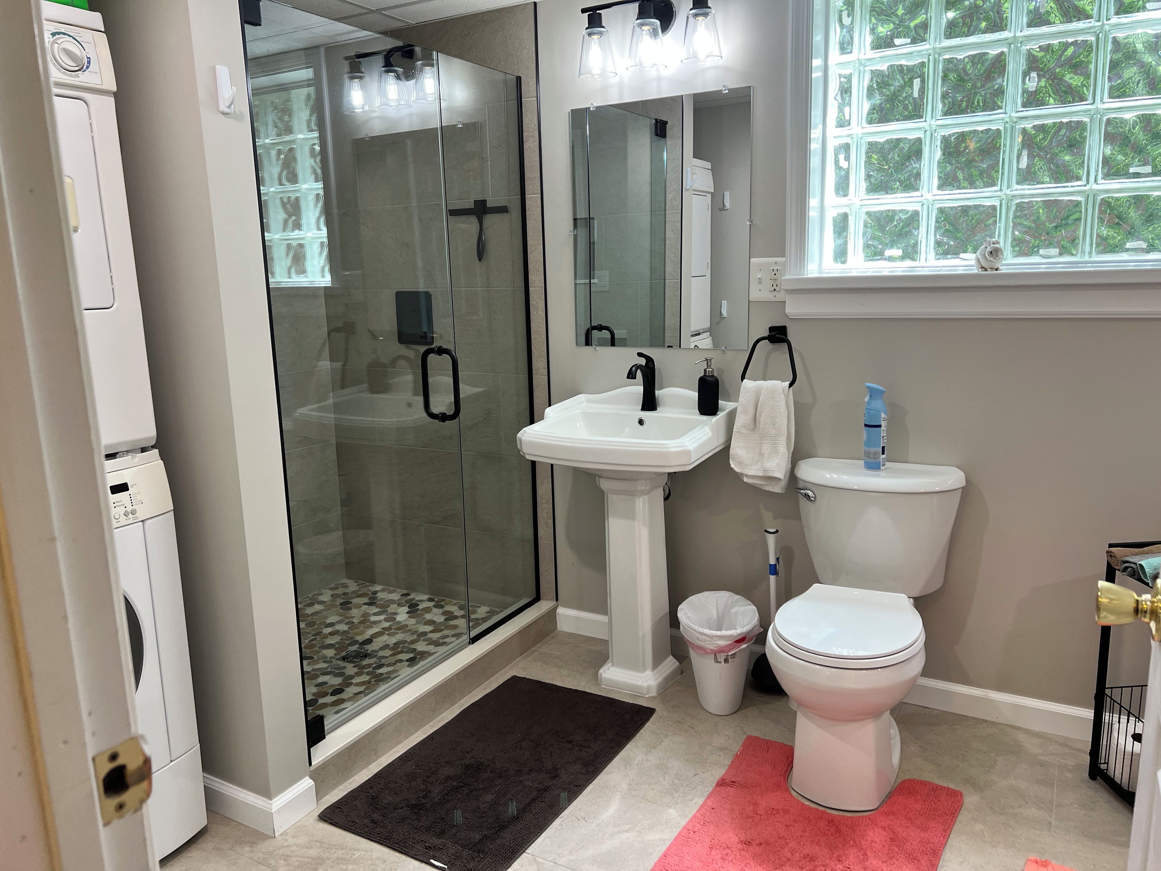 A modern bathroom with a glass-enclosed shower, a pedestal sink, a toilet, and a window with a view of greenery.