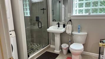 A modern bathroom with a glass-enclosed shower, a pedestal sink, a toilet, and a window with a view of greenery.