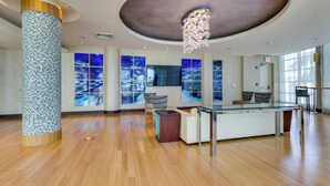A spacious lobby with a wooden floor, a large pillar with mosaic tiles, a glass-top reception desk, and a chandelier.