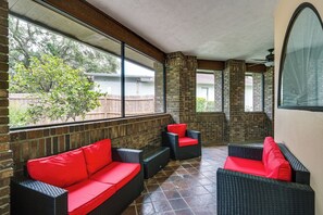 Een overdekt terras met rieten meubels en rode kussens, grote ramen en bakstenen pilaren.
