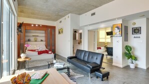 Uma sala de estar moderna com sofá, mesa de centro e uma mesa de jantar com petiscos. Há uma cama com manta vermelha e uma planta em vaso.