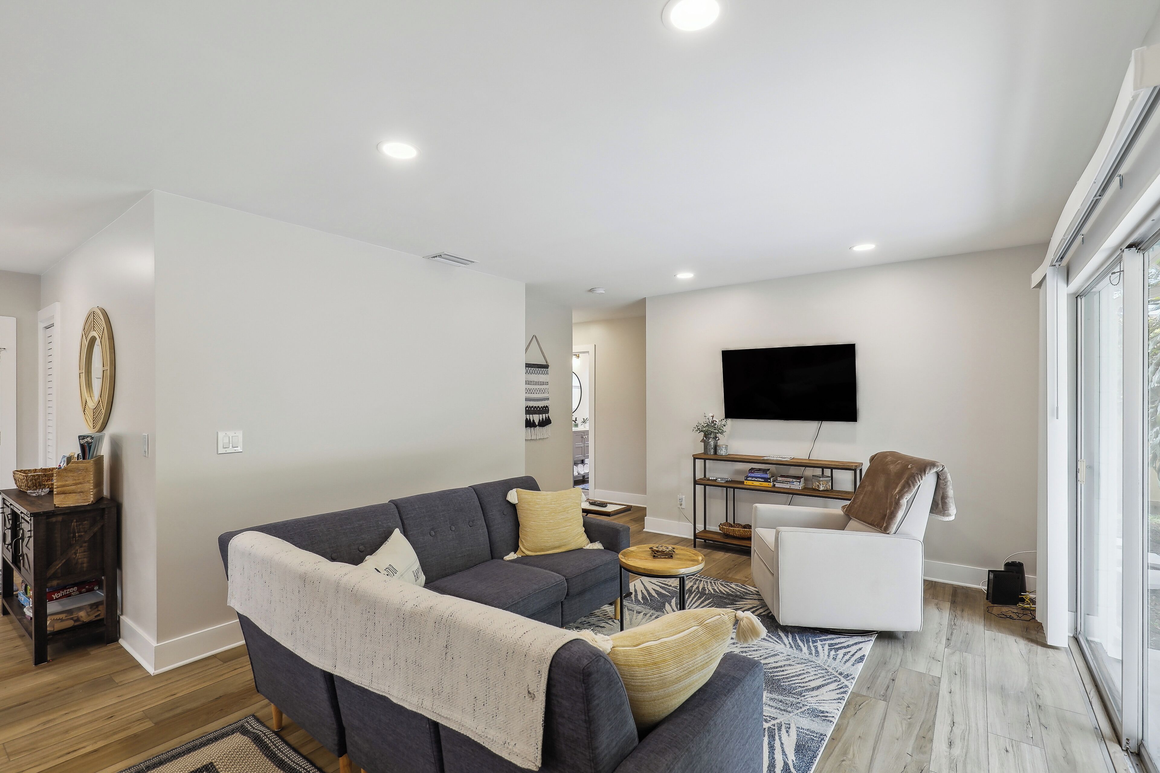 A modern living room with a grey sofa, a white armchair, a small round coffee table, and a flat-screen TV mounted on the wall.
