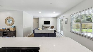 Una cocina moderna con encimera blanca, estufa integrada y un rincón de comedor con sofá azul oscuro y televisor al fondo.