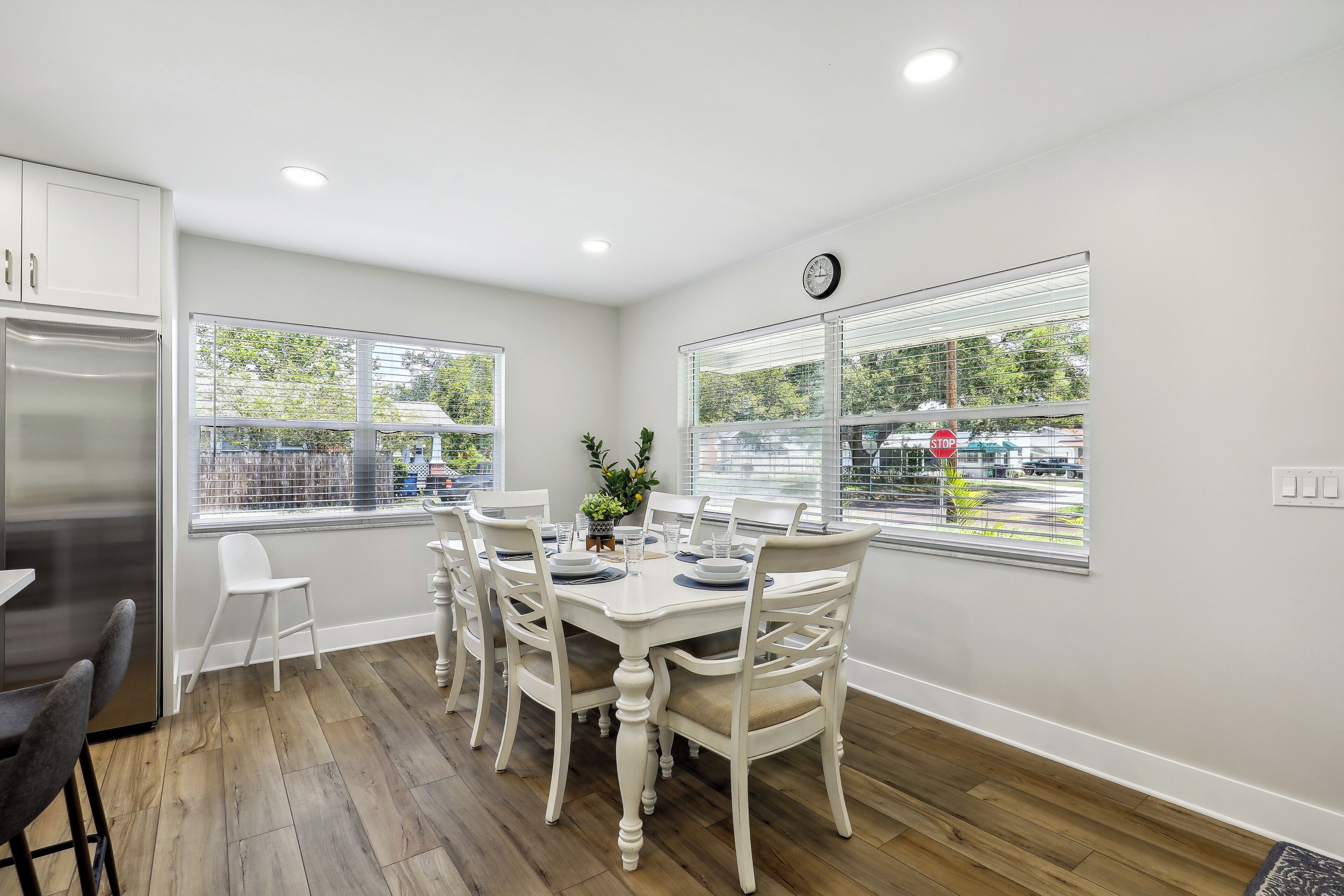A dining area with a white table and chairs, a clock on the wall, and windows with blinds.