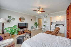 A modern living room with a sofa, a coffee table, a TV, and a ceiling fan. There is a kitchen area with cabinets and a refrigerator.