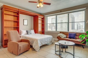 A bedroom with a bed, a chair, a sofa, a small table, and a large window with blinds.
