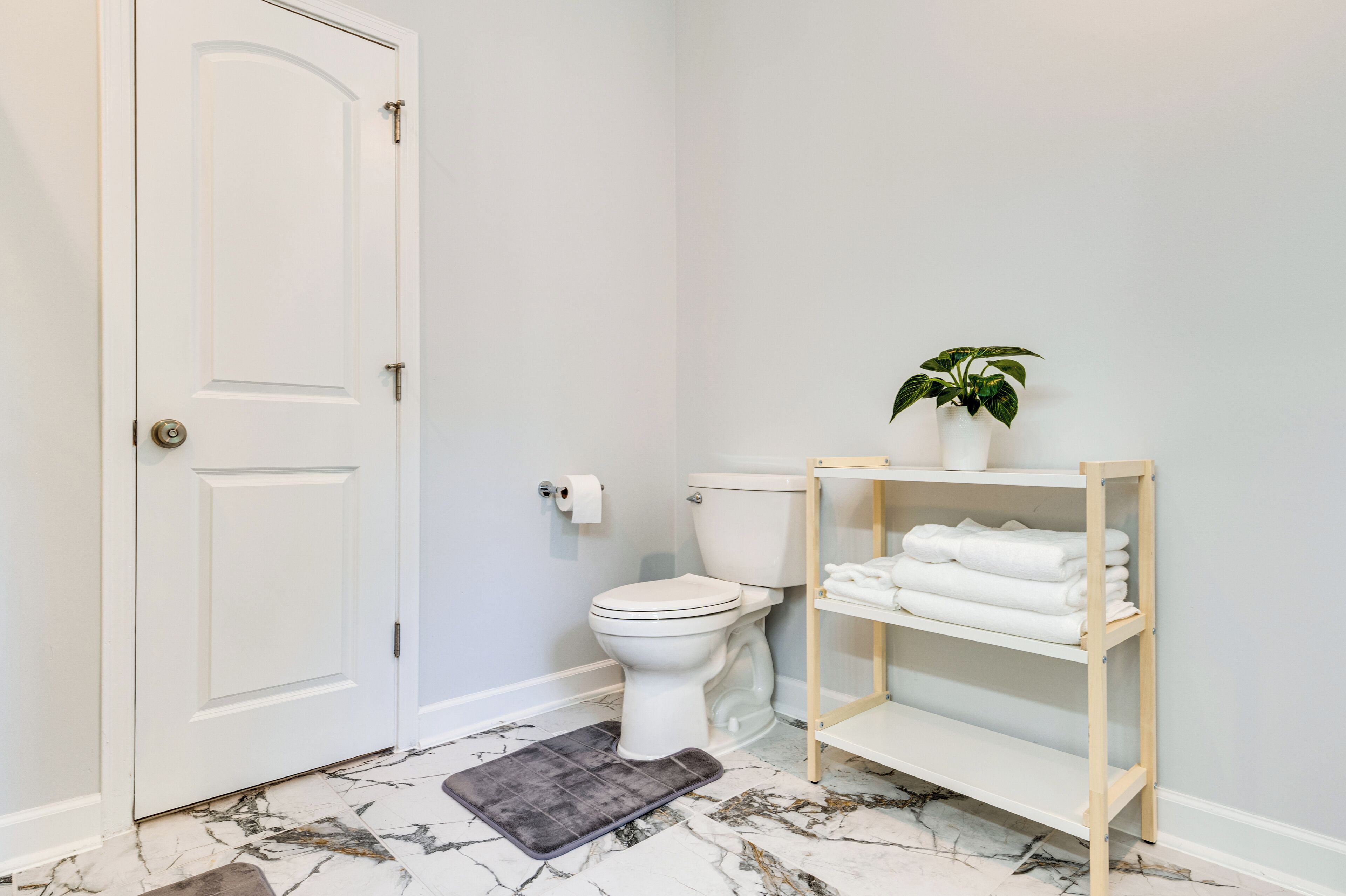 Une salle de bain avec des toilettes, une porte blanche, un sol en marbre, un petit tapis, une étagère avec des serviettes et une plante en pot.