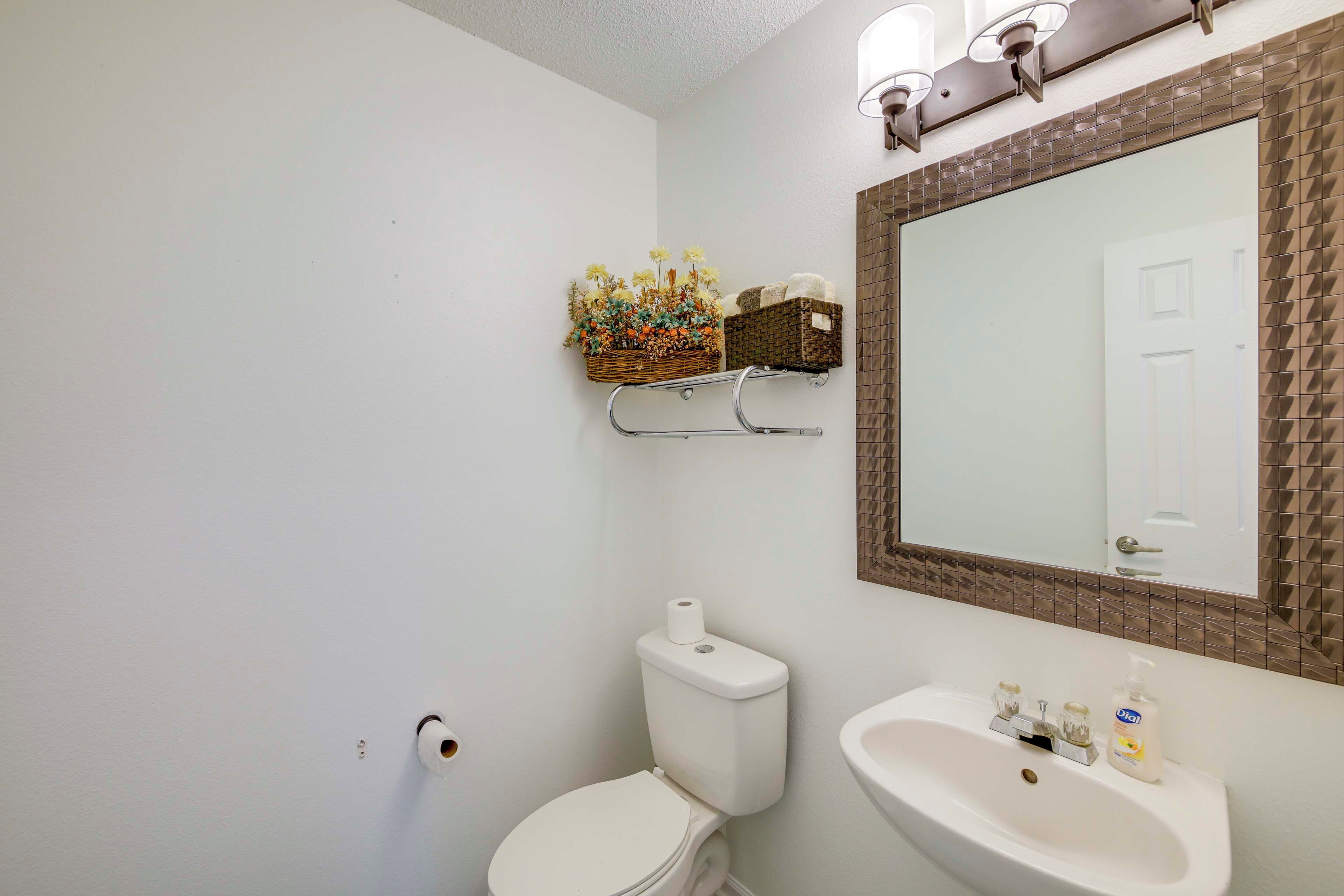 Une salle de bain avec des toilettes, un lavabo et un miroir doté d’un cadre marron. Un panier rempli de fleurs est accroché au mur, et un rouleau de papier toilette se trouve sur les toilettes.