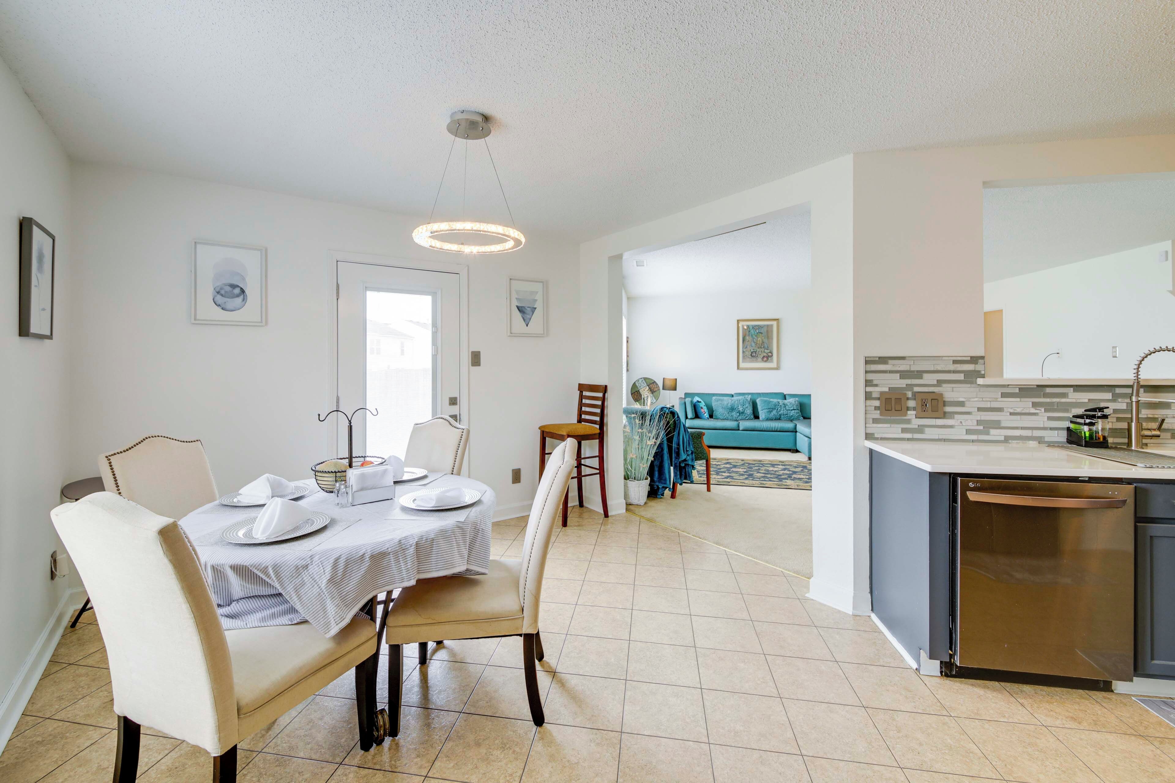 Uma cozinha moderna com lava-louças, sala de jantar com cadeiras e mesa posta, e sala de estar com sofá e poltronas.