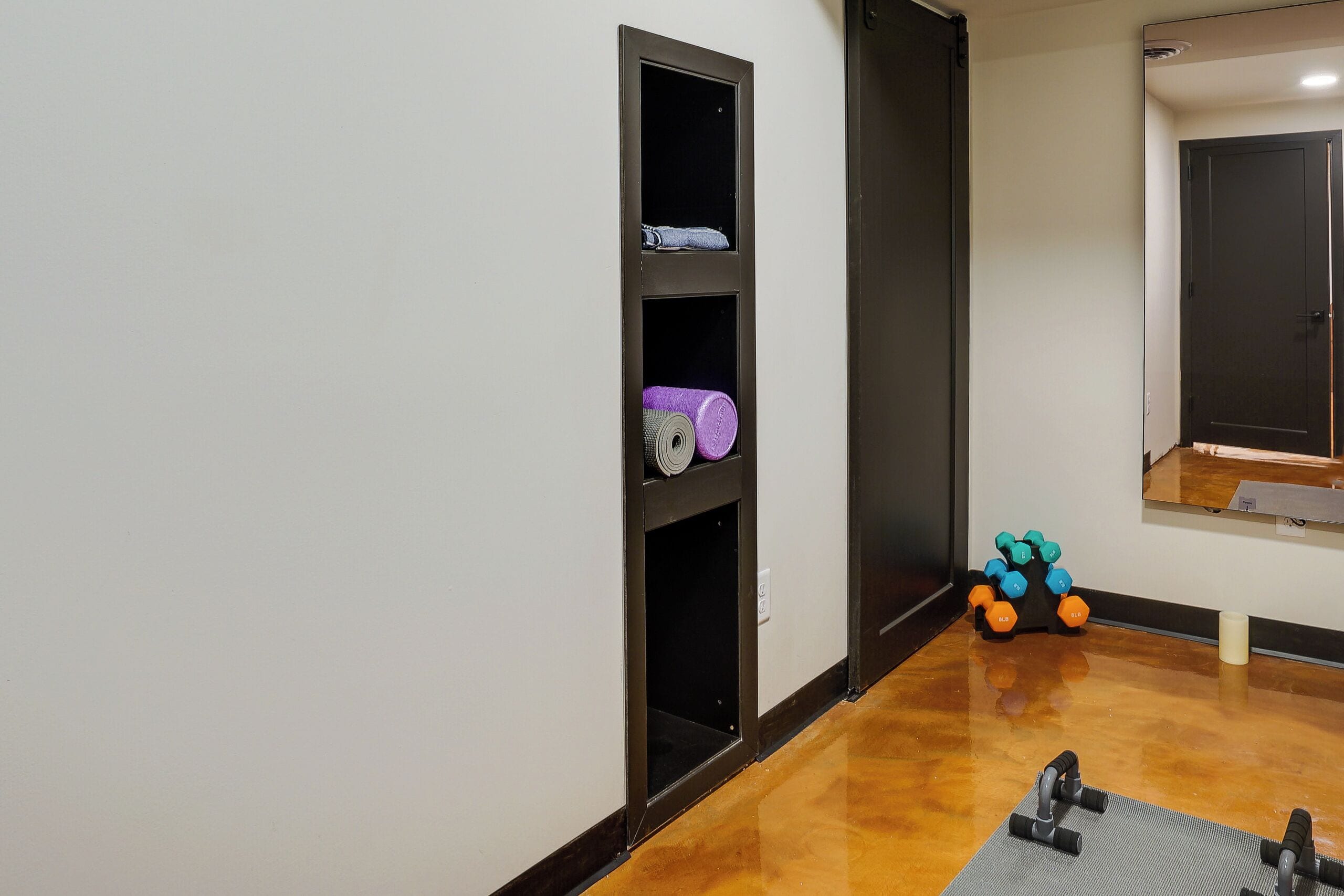 A hallway with a mirrored wall, a shelf with rolled-up items, and a pair of dumbbells on the floor.