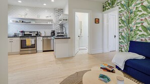 Una cocina moderna con armarios blancos y suelo de madera, que da paso a una sala de estar con sofá azul y una mesa de centro.