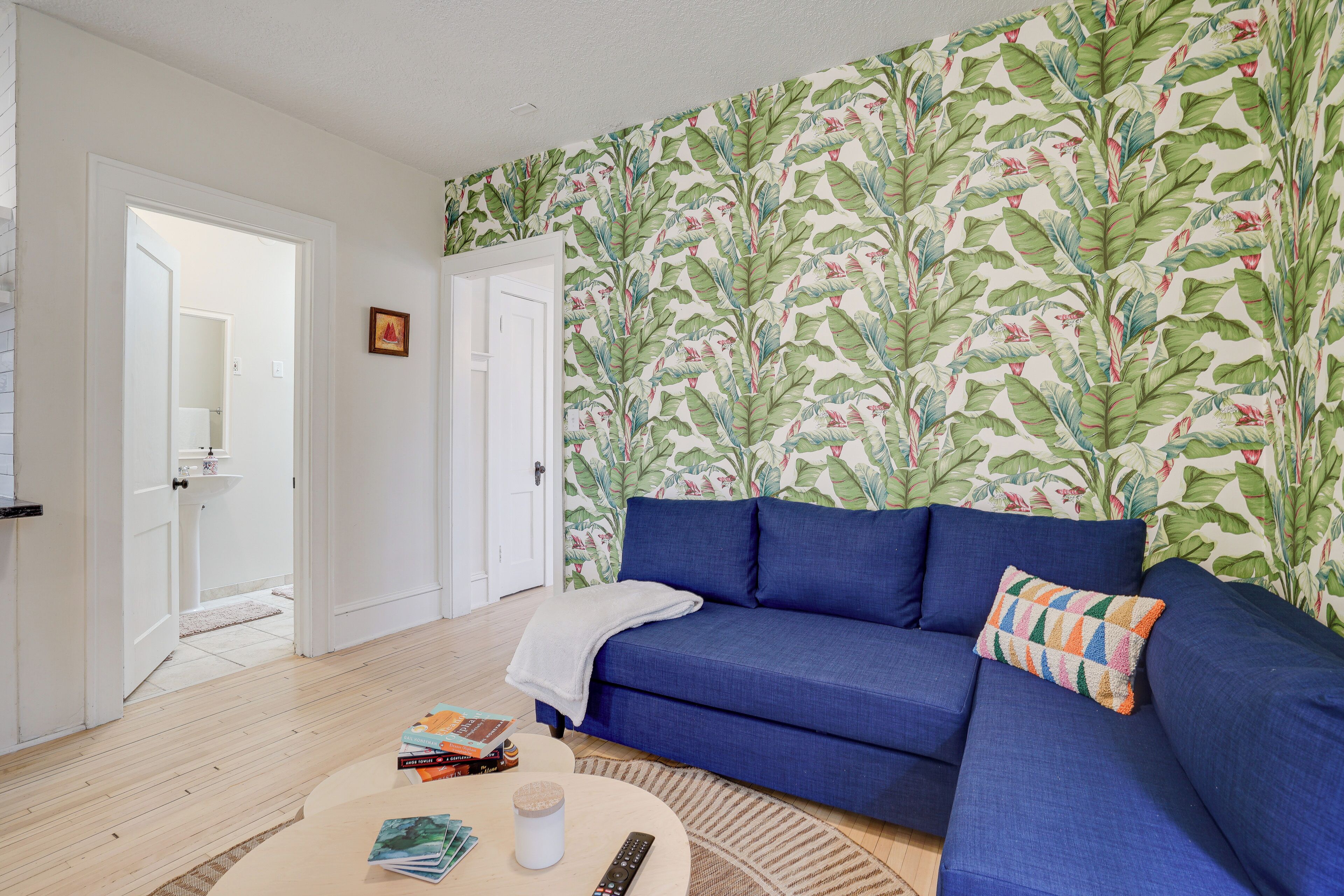 A living room with a blue sofa, a coffee table with books and a remote, and a wall with a tropical leaf pattern.